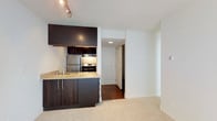 This single-floor residence offers a clean, modern aesthetic defined by thoughtful design and quality finishes throughout. The open-concept layout seamlessly connects the kitchen and living area, creating a functional and cohesive living space. The kitchen is outfitted with dark espresso cabinetry that extends from floor to ceiling, providing ample storage, while stainless steel appliances — including a refrigerator, gas range, and integrated range hood — add a polished, contemporary touch. A granite-topped counter with an undermount sink completes the kitchen, offering both style and practicality. Track lighting overhead ensures the space is well-illuminated. The adjoining living area features plush carpeting in a neutral tone, contributing to a warm and inviting ambiance. Rich hardwood flooring transitions into the hallway, which leads to additional areas of the unit, including a closet and a paneled entry door that adds a classic architectural detail. The crisp white walls and clean lines throughout lend the space a bright, airy feel. Overall, this unit combines modern finishes with a practical layout, making it a comfortable and stylish living environment.