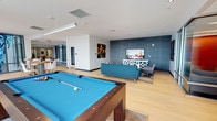 This community game room is a well-appointed social space designed to foster leisure and connection. The room's contemporary design is immediately apparent, featuring clean lines, a bright white ceiling with recessed and linear lighting, and light hardwood-style flooring that gives the space a fresh, open feel. A bold blue accent wall anchors the lounge area, creating a striking focal point that complements the room's modern aesthetic. A full-size billiards table with a vibrant blue felt surface takes center stage, serving as the room's primary recreational feature and inviting friendly competition. Adjacent to the billiards area, a comfortable lounge zone is furnished with a teal sofa, accent chairs, and a stylish geometric media console, all arranged around a large flat-screen television — ideal for watching sports or hosting casual gatherings. Colorful artwork adorns the walls, adding personality and energy to the space. The open layout allows for easy movement throughout the room, accommodating multiple activities simultaneously. With its thoughtful combination of recreational amenities, inviting seating, and vibrant design elements, this community game room offers residents a dynamic and engaging space to relax and socialize.
