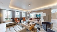 Located at 1881 N Rollins Rd in Burlingame, California, this sophisticated single-floor community space offers a generously proportioned open-concept layout designed with a refined, contemporary aesthetic. The interior is anchored by a spacious lounge area featuring coffered ceilings with warm ambient cove lighting, an elegant triangular pendant light fixture, and large floor-to-ceiling windows that flood the space with natural light while framing views of the surrounding landscape.

The lounge is furnished with a curated mix of plush sofas, tufted benches, and cognac leather wingback chairs arranged around a substantial wood coffee table, all set atop a patterned area rug. A dedicated seating alcove with built-in shelving and a mounted display screen provides an additional gathering zone, complemented by warm-toned wall sconces and a striking stone accent feature.

Transitioning seamlessly through an arched passageway, the kitchen and dining area showcases large-format marble tile flooring, dual oversized islands with sleek stone countertops, professional-grade stainless steel appliances, and a row of bold brass globe pendant lights. A formal dining table with upholstered chairs sits adjacent to a teal-accented bar area, completing this well-appointed communal space. Throughout, the design balances warm neutrals with luxurious material finishes, creating an inviting and polished atmosphere.