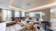 Located at 1881 N Rollins Rd in Burlingame, California, this sophisticated single-floor community space offers a generously proportioned open-concept layout designed with a refined, contemporary aesthetic. The interior is anchored by a spacious lounge area featuring coffered ceilings with warm ambient cove lighting, an elegant triangular pendant light fixture, and large floor-to-ceiling windows that flood the space with natural light while framing views of the surrounding landscape.

The lounge is furnished with a curated mix of plush sofas, tufted benches, and cognac leather wingback chairs arranged around a substantial wood coffee table, all set atop a patterned area rug. A dedicated seating alcove with built-in shelving and a mounted display screen provides an additional gathering zone, complemented by warm-toned wall sconces and a striking stone accent feature.

Transitioning seamlessly through an arched passageway, the kitchen and dining area showcases large-format marble tile flooring, dual oversized islands with sleek stone countertops, professional-grade stainless steel appliances, and a row of bold brass globe pendant lights. A formal dining table with upholstered chairs sits adjacent to a teal-accented bar area, completing this well-appointed communal space. Throughout, the design balances warm neutrals with luxurious material finishes, creating an inviting and polished atmosphere.