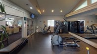 This single-floor fitness center offers a comprehensive workout environment designed for both cardio and strength training. The space is outfitted with a range of equipment, including elliptical machines, a stationary recumbent bike, a multi-station cable machine, and additional cardio options, all arranged efficiently across a durable dark rubber flooring. Large wall-mounted mirrors line one side of the room, amplifying the sense of space and allowing users to monitor their form during exercise. Recessed lighting throughout the ceiling provides consistent, even illumination, while multiple windows along the walls invite generous natural light and views of the lush, green surroundings outside. A glass-panel door provides direct access to an outdoor pool area, seamlessly connecting indoor and outdoor amenities. A mounted flat-screen television is positioned for visibility across the workout floor. The entry area features a glass-front display case and informational signage, including posted exercise room rules and an emergency phone. Adjacent to the fitness room, a comfortable lounge area with seating and a fireplace is visible, offering a relaxing space to unwind before or after a workout. A tall decorative plant near the entrance adds a welcoming, natural touch to the overall atmosphere.