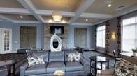 This well-appointed single-floor space exudes a refined, contemporary aesthetic defined by thoughtful architectural details and upscale finishes. The centerpiece of the main lounge area is a striking stacked-stone fireplace wall, which anchors the room and provides a warm, textural contrast to the smooth, painted surfaces surrounding it. A flat-screen television is mounted above the fireplace, seamlessly integrating entertainment into the elegant design.

The ceiling draws immediate attention with its distinctive coffered design, featuring recessed panels framed by crisp white molding. A large drum pendant light hangs centrally, complementing the recessed lighting embedded throughout the ceiling grid and contributing to a layered, well-balanced lighting scheme.

The walls are finished in a cool blue-gray palette that lends the space a calm and sophisticated atmosphere. Decorative wall panels flank the fireplace, adding visual depth and artistic character. Large windows dressed with patterned drapes allow natural light to filter into the room while maintaining a sense of privacy.

The seating area is anchored by a substantial charcoal leather sectional sofa adorned with floral accent pillows, paired with additional seating to create an inviting, social arrangement. Dark hardwood flooring and an area rug ground the space, while a sculptural ceramic vase serves as a striking decorative focal point. Overall, the space combines comfort with upscale design sensibility.
