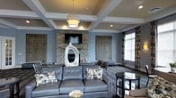 This well-appointed single-floor space exudes a refined, contemporary aesthetic defined by thoughtful architectural details and upscale finishes. The centerpiece of the main lounge area is a striking stacked-stone fireplace wall, which anchors the room and provides a warm, textural contrast to the smooth, painted surfaces surrounding it. A flat-screen television is mounted above the fireplace, seamlessly integrating entertainment into the elegant design.

The ceiling draws immediate attention with its distinctive coffered design, featuring recessed panels framed by crisp white molding. A large drum pendant light hangs centrally, complementing the recessed lighting embedded throughout the ceiling grid and contributing to a layered, well-balanced lighting scheme.

The walls are finished in a cool blue-gray palette that lends the space a calm and sophisticated atmosphere. Decorative wall panels flank the fireplace, adding visual depth and artistic character. Large windows dressed with patterned drapes allow natural light to filter into the room while maintaining a sense of privacy.

The seating area is anchored by a substantial charcoal leather sectional sofa adorned with floral accent pillows, paired with additional seating to create an inviting, social arrangement. Dark hardwood flooring and an area rug ground the space, while a sculptural ceramic vase serves as a striking decorative focal point. Overall, the space combines comfort with upscale design sensibility.