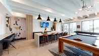 This single-floor amenity space is designed with a modern aesthetic, combining social and recreational functions in an open, well-appointed layout. Exposed beam ceilings finished in white lend architectural character throughout, while large-format tile flooring unifies the space and contributes to its clean, contemporary feel.

The game and social area serves as a central focal point, anchored by a solid wood billiards table set atop a bold geometric patterned area rug. Nearby, a long bar-height counter with warm wood barstools provides casual seating for multiple guests, illuminated overhead by a trio of striking black industrial pendant lights. A wall-mounted flat-screen television is positioned above the counter for entertainment.

Adjacent to the game area, a comfortable lounge zone features plush seating, leather accent chairs, and a substantial wood coffee table. Floor-to-ceiling windows and black-framed glass doors flood this area with natural light and offer direct access to an outdoor terrace, providing a seamless indoor-outdoor connection. A wall-mounted television and a warm wood credenza complete the lounge furnishings.

Decorative artwork and sculptural accents are thoughtfully placed throughout, contributing to the space's curated, upscale ambiance. A separate enclosed room, visible through glass partitions, offers additional gathering space.