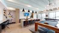 This single-floor amenity space is designed with a modern aesthetic, combining social and recreational functions in an open, well-appointed layout. Exposed beam ceilings finished in white lend architectural character throughout, while large-format tile flooring unifies the space and contributes to its clean, contemporary feel.

The game and social area serves as a central focal point, anchored by a solid wood billiards table set atop a bold geometric patterned area rug. Nearby, a long bar-height counter with warm wood barstools provides casual seating for multiple guests, illuminated overhead by a trio of striking black industrial pendant lights. A wall-mounted flat-screen television is positioned above the counter for entertainment.

Adjacent to the game area, a comfortable lounge zone features plush seating, leather accent chairs, and a substantial wood coffee table. Floor-to-ceiling windows and black-framed glass doors flood this area with natural light and offer direct access to an outdoor terrace, providing a seamless indoor-outdoor connection. A wall-mounted television and a warm wood credenza complete the lounge furnishings.

Decorative artwork and sculptural accents are thoughtfully placed throughout, contributing to the space's curated, upscale ambiance. A separate enclosed room, visible through glass partitions, offers additional gathering space.