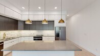 This thoughtfully designed single-floor residence showcases a clean, contemporary aesthetic throughout. The centerpiece of the home is a striking chef's kitchen, appointed with two-tone cabinetry—crisp white upper cabinets paired with dark wood lower cabinets—set against a marble-inspired tile backsplash. A generous island with a sleek grey quartz countertop anchors the space, illuminated by a trio of brushed gold dome pendant lights. Stainless steel appliances, including a French door refrigerator and range, complete the culinary space.

The open living area is defined by a dramatic floor-to-ceiling black-framed glass wall with multi-panel sliding doors, flooding the room with natural light and offering direct access to an outdoor area with views of surrounding greenery. Light oak hardwood flooring flows consistently throughout the main living areas and bedrooms, enhancing the bright, airy atmosphere.

Bedrooms are well-proportioned and feature modern sliding panel doors, with one offering a spacious walk-in closet fitted with wire shelving and hanging rods. Two bathrooms serve the residence: one features a glass-enclosed walk-in shower with white subway tile and brushed gold fixtures, while the second includes a full tub-shower combination with a curtain rod and matching gold hardware, both finished with dark grey tile flooring.