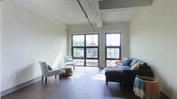 Situated on North High Street in Columbus, Ohio's vibrant Short North neighborhood, this single-floor space blends industrial character with modern comfort. The open living area immediately draws attention with its soaring ceilings accented by exposed structural beams and overhead piping, lending an authentic loft-like aesthetic. Rich, dark hardwood flooring runs throughout, providing a warm contrast to the light-toned walls. A striking set of large, black-framed windows dominates the exterior wall, inviting generous amounts of natural light to fill the space and offering views of the surrounding neighborhood. The windows also provide access to or frame an outdoor area, further enhancing the sense of openness. The layout is versatile and well-proportioned, comfortably accommodating distinct seating arrangements that make the space feel both functional and inviting. The overall ambiance strikes a balance between urban industrial design and a welcoming, livable atmosphere, making it well-suited for a variety of uses. Its prime location along one of Columbus's most well-known commercial and cultural corridors adds significant appeal for those seeking a dynamic urban environment.
