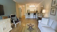 This single-story residence showcases an open-concept layout that seamlessly connects the living area to the kitchen, creating a spacious and cohesive environment. Warm hardwood floors extend throughout the main living space, complemented by crown molding that adds a refined architectural detail to the ceilings. The living area is well-proportioned, accommodating a comfortable seating arrangement that includes a classic upholstered sofa and an ornate accent chair, centered around a decorative coffee table atop a patterned area rug. A central column subtly defines the transition between the living and kitchen zones without interrupting the open flow. The kitchen features white cabinetry, stainless steel appliances, and a wraparound breakfast bar with dark bar stools, providing both functional prep space and casual dining. Pendant lighting above the kitchen area adds focused illumination and a stylish touch. Neutral wall tones in soft green and beige create a calming backdrop throughout. A doorway visible in the background suggests additional rooms beyond the main living space. Overall, the layout balances functionality with an approachable, welcoming ambiance.