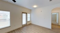 This single-floor space offers a clean, minimalist design with a neutral color palette throughout. The room features light hardwood-style flooring that runs seamlessly across the open area, complementing the crisp white walls and creating a bright, airy atmosphere. A glass-paneled door, fitted with horizontal blinds, provides direct access to the outdoors while allowing natural light to fill the interior. An adjacent window with matching blinds further enhances the room's brightness and ventilation options. The ceiling is smooth and white, accented by a flush-mounted light fixture and a ceiling fan visible in the corner, ensuring year-round comfort. Multiple electrical outlets are positioned at convenient intervals along the walls, offering practical utility throughout the space. The overall design is straightforward and functional, with a well-maintained, move-in-ready condition. The light tones of the flooring and walls work together to maximize the sense of openness, making the space feel welcoming and versatile for a variety of uses.