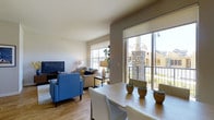 This single-floor residence showcases a thoughtfully designed open-concept layout where the dining and living areas flow together naturally, creating a cohesive and functional living environment. The space is bathed in natural light, thanks to large windows fitted with roller shades and a sliding glass door that opens to a private balcony overlooking a stone-accented exterior courtyard area.

The living area is warm and inviting, centered around a flat-screen television and complemented by a comfortable seating arrangement featuring a bold blue accent chair and a neutral-toned sofa. Light hardwood flooring runs throughout, lending warmth and continuity to the space. A coordinating area rug anchors the living zone, adding texture and definition.

The dining area sits adjacent to the living space, furnished with a light wood dining table and upholstered chairs. Decorative succulents in terracotta planters serve as a simple, tasteful centerpiece. The neutral wall tones throughout create a calm, versatile backdrop that enhances the overall sense of openness. Together, the abundant natural light, outdoor access, and cohesive design make this a comfortable and well-appointed single-level home.