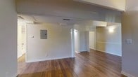This single-floor space is characterized by its open layout and warm hardwood flooring that extends throughout the main living area. Structural beams are visible along the ceiling, adding an architectural element that defines the space overhead. A decorative column stands near the center of the room, subtly separating zones while maintaining an open feel. The walls are finished in a clean, neutral white, providing a bright and versatile backdrop. An electrical panel is mounted on one wall, and HVAC vents are integrated into the ceiling for climate control. Beyond the main area, a lit hallway leads to additional rooms, including what appears to be a closeted space and a doorway to further interior rooms. The layout suggests a functional flow between areas. Overall, the space offers a straightforward, unfurnished interior with classic hardwood floors and a neutral palette, making it highly adaptable for a variety of uses.