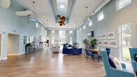 Located at 500 Waters Edge Dr in Newport News, Virginia, this single-floor leasing and community office for Compass at City Center offers a well-appointed, open layout designed to welcome and inform prospective residents. The space is defined by soaring ceilings with clerestory windows positioned near the roofline, flooding the interiors with abundant natural light and creating an airy, expansive atmosphere throughout.

The reception area greets visitors with a clean, modern front desk, warm wood-look flooring, and soft blue-grey walls that establish a calm, professional tone. Decorative wall art and lush potted plants add warmth to the space. Double glass doors and a connecting corridor provide access to additional rooms and offices.

The combined kitchen and living area serves as the heart of the space. The kitchen features floor-to-ceiling white shaker cabinetry, quartz countertops, a mosaic tile backsplash, stainless steel appliances, and a large island with seating — all illuminated by pendant lighting and surrounded by large windows with outdoor views.

The adjacent lounge area is furnished with teal and navy seating, round dining tables, and a wall-mounted television, creating a comfortable, community-oriented setting. The cohesive coastal color palette, hardwood-style flooring, and abundant natural light give the entire space a polished yet inviting character.