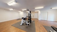 Located at 1500 Coastal Lane in Myrtle Beach, SC, this single-floor fitness center is designed to support a well-rounded exercise routine in a clean, functional environment. The space features warm wood-style flooring throughout, complemented by bright fluorescent lighting that ensures the entire area is evenly and thoroughly illuminated. Protective rubber floor mats are strategically placed beneath equipment stations, adding both safety and structure to the layout.

The cardio section is well-equipped with a NordicTrack treadmill, a rowing machine, an elliptical trainer, and a recumbent stationary bike, providing a comprehensive range of aerobic workout options. Strength training is supported by a multi-station cable and weight machine, suitable for targeting multiple muscle groups. Additional open floor space with foam mats offers room for stretching, bodyweight exercises, or yoga.

The facility includes multiple entry and exit points, clearly marked with illuminated exit signs, as well as a water fountain station for convenient hydration. Windows along the perimeter allow natural light to filter in, contributing to an open and welcoming atmosphere. A louvered storage closet and additional utility doors round out the practical amenities of this well-appointed fitness space.
