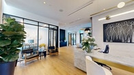 Located at 777 Northwest 2nd Avenue in Miami, FL, this single-floor commercial space delivers a refined, contemporary aesthetic across a well-appointed layout. Light oak hardwood flooring runs throughout, anchoring a cohesive design that balances openness with defined functional zones.

The welcoming reception area centers on a striking marble desk with gold accents, paired with cream upholstered chairs and an elegant sputnik-style pendant light. Floor-to-ceiling sheer curtains filter abundant natural light from expansive windows, creating a bright and airy atmosphere.

The living/lounge area features a bold navy textured accent wall alongside a sleek credenza, offset by a dramatic drum-shaped pendant chandelier overhead. Curved sofas and accent chairs form an inviting seating arrangement, while lush indoor plants add organic warmth throughout the space.

Glass-walled, black-framed office enclosures provide privacy without sacrificing visual connectivity. One office is furnished with a dark marble-top conference table and cognac leather chairs, accented by vibrant curated artwork. A second private office accommodates a dedicated workstation with yellow accent seating.

The hallway transitions seamlessly between zones, maintaining the space's clean, modern flow. Every detail — from the sculptural light fixtures to the carefully selected art pieces — reflects a polished, design-forward sensibility suited to a premium professional environment.