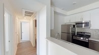 This contemporary single-floor residence is designed with a clean, modern aesthetic throughout. The open-concept kitchen serves as a central feature, showcasing crisp white cabinetry, a smooth white countertop with a peninsula that doubles as a breakfast bar, and a full suite of stainless steel appliances including a refrigerator, microwave, and gas range. A tiled backsplash adds a subtle decorative touch against the otherwise minimalist color palette. The kitchen flows seamlessly into the surrounding living areas, creating an open and airy atmosphere. Light wood-tone flooring runs throughout, complementing the bright white walls and ceilings that maximize the sense of space. A well-lit hallway extends from the kitchen area, lined with flush doors and a ceiling-mounted HVAC vent, leading toward a bathroom visible at the far end. Recessed and flush-mount lighting ensures consistent illumination across the unit. The overall design emphasizes functionality and simplicity, with clean lines and a neutral palette that creates a welcoming, move-in-ready environment well-suited for modern living.