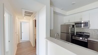 This contemporary single-floor residence is designed with a clean, modern aesthetic throughout. The open-concept kitchen serves as a central feature, showcasing crisp white cabinetry, a smooth white countertop with a peninsula that doubles as a breakfast bar, and a full suite of stainless steel appliances including a refrigerator, microwave, and gas range. A tiled backsplash adds a subtle decorative touch against the otherwise minimalist color palette. The kitchen flows seamlessly into the surrounding living areas, creating an open and airy atmosphere. Light wood-tone flooring runs throughout, complementing the bright white walls and ceilings that maximize the sense of space. A well-lit hallway extends from the kitchen area, lined with flush doors and a ceiling-mounted HVAC vent, leading toward a bathroom visible at the far end. Recessed and flush-mount lighting ensures consistent illumination across the unit. The overall design emphasizes functionality and simplicity, with clean lines and a neutral palette that creates a welcoming, move-in-ready environment well-suited for modern living.