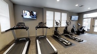 This fitness center is a dedicated, single-floor exercise facility designed to provide residents or members with a convenient and comfortable workout environment. The space is outfitted with a lineup of modern cardio equipment, including multiple treadmills and elliptical machines, all arranged to maximize usable floor space while maintaining an open, uncluttered feel. Protective floor mats are placed beneath the equipment, reflecting a thoughtful approach to both safety and maintenance. Natural light enters through a row of windows fitted with white blinds, creating a bright and welcoming atmosphere. Two wall-mounted flat-screen televisions are positioned at either end of the room, offering entertainment to keep users engaged during their workouts. The walls are finished in a neutral gray tone that contributes to a clean, modern aesthetic, while the carpeted flooring adds a subtle warmth to the space. Recessed ceiling lighting supplements the natural light, ensuring consistent illumination throughout. The overall design is functional and practical, striking a balance between a professional gym setting and an accessible, community-oriented fitness space.
