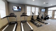 This fitness center is a dedicated, single-floor exercise facility designed to provide residents or members with a convenient and comfortable workout environment. The space is outfitted with a lineup of modern cardio equipment, including multiple treadmills and elliptical machines, all arranged to maximize usable floor space while maintaining an open, uncluttered feel. Protective floor mats are placed beneath the equipment, reflecting a thoughtful approach to both safety and maintenance. Natural light enters through a row of windows fitted with white blinds, creating a bright and welcoming atmosphere. Two wall-mounted flat-screen televisions are positioned at either end of the room, offering entertainment to keep users engaged during their workouts. The walls are finished in a neutral gray tone that contributes to a clean, modern aesthetic, while the carpeted flooring adds a subtle warmth to the space. Recessed ceiling lighting supplements the natural light, ensuring consistent illumination throughout. The overall design is functional and practical, striking a balance between a professional gym setting and an accessible, community-oriented fitness space.