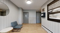 This single-floor lobby serves as the primary entrance and circulation hub of a modern residential building. The space is defined by its consistent Scandinavian-inspired aesthetic, featuring light wood-look flooring and vertical shiplap wall paneling in soft grey tones throughout, creating a cohesive and contemporary atmosphere.

The main entryway is anchored by full-height glass double doors framed in matte black, allowing an abundance of natural light to flood the interior and offering clear sightlines to the landscaped exterior. A large entry mat leads visitors inward, where a comfortable grey accent chair, a round side table, and a potted palm contribute a warm, welcoming ambiance.

The lobby transitions into a secondary vestibule area equipped with an elevator and staircase providing access to upper-level residential units, including a distinctively teal-colored unit door visible along the corridor. This area also houses a wall-mounted fire alarm control panel and intercom system, underscoring the building's safety infrastructure.

Circular flush-mounted ceiling fixtures provide consistent, even illumination throughout. A round decorative mirror and framed wall art add thoughtful design touches, while the overall layout remains open and uncluttered. The combination of functional building systems and refined finishes makes this lobby both practical and visually appealing.