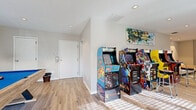 This dedicated game room delivers an impressive recreational experience within a thoughtfully designed single-floor layout. Light wood-look flooring runs throughout the space, lending a clean, modern foundation that contrasts playfully with the colorful entertainment elements on display. The room is organized into distinct zones, beginning with an arcade area showcasing a collection of full-size classic cabinets, including Ms. Pac-Man, Mortal Kombat II, and NBA Jam, positioned beneath angled ceilings with recessed lighting. A vibrant abstract canvas adorns the wall above, adding an artistic touch to the gaming corner. Transitioning deeper into the space, a stylish lounge area features a tufted sofa set against a striking textured accent wall with a chevron-patterned design, providing a comfortable retreat between games. The main open area is anchored by a contemporary pool table with a bold blue felt surface, bathed in natural light streaming through large windows and glass doors that connect to the outdoors. Recessed ceiling lights illuminate the entire space evenly, while colorful area rugs define each zone. A bookshelf display, framed artwork, and decorative accessories throughout contribute to the space's dynamic yet welcoming character.