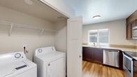 This single-floor residence offers a functional and comfortable layout with a clean, neutral aesthetic throughout. The living area features warm hardwood-style flooring and a textured ceiling, with a sliding glass door that invites natural light and provides direct access to an outdoor patio area. The kitchen is well-equipped with rich dark-stained cabinetry, light countertops, a double-basin stainless steel sink positioned beneath a window overlooking the exterior, and a stainless steel dishwasher. Ample upper and lower cabinet storage makes this an efficient workspace. A dedicated in-unit laundry area includes a full-size washer and dryer set with overhead wire shelving for additional storage convenience. The bathroom presents a clean, modern design with a dark wood vanity, white countertop, a large mirror, tiled shower surround, and light hardwood-style flooring. Bedrooms feature plush carpeting, closet access, and a calm, neutral color palette. A hallway connects the living spaces smoothly, with hardwood-style flooring continuing through the common areas. The overall ambiance is bright, practical, and move-in ready, offering a well-organized layout suited for comfortable everyday living.