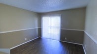 This single-floor space offers a clean and functional layout designed for comfortable everyday living. The main living area showcases rich, dark hardwood flooring that adds warmth and a polished look throughout. Walls are finished in a neutral taupe tone and feature a distinctive two-tone treatment, divided by a horizontal chair rail that adds a subtle architectural detail. White baseboards provide a crisp contrast, enhancing the overall tidiness of the space. A textured popcorn ceiling adds a classic character overhead. The standout feature of the room is a full-width sliding glass door dressed with vertical blinds, which serves as both a functional entry point to the outdoors and a generous source of natural light. When the blinds are drawn open, the room is bathed in bright, diffused daylight, creating an airy and inviting atmosphere. The layout is open and versatile, providing ample room for a variety of furniture arrangements. The combination of neutral tones, natural light, and durable flooring makes this a practical and welcoming space suited for a range of uses.