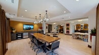 The Drawing Room, located in Houston's 77008 zip code, is a thoughtfully designed single-floor communal space that blends professional functionality with inviting social ambiance. The layout is open and airy, anchored by a striking live-edge wood conference table surrounded by ergonomic leather chairs and illuminated by a trio of geometric pendant lights. Floor-to-ceiling sliding glass doors line the perimeter, flooding the space with natural light and offering views of lush, tree-lined outdoor surroundings. Light hardwood flooring runs throughout the main area, enhancing the bright and welcoming atmosphere. A dedicated media lounge features pairs of teal velvet accent chairs arranged around a geometric gold coffee table, with a wall-mounted flat-screen TV and a built-in dark cabinetry unit displaying books and décor beneath a curated gallery of framed black-and-white photographs. Bold golden-yellow accent walls punctuate the space and provide a warm contrast to the neutral tones. Additional seating nooks with round tables and upholstered chairs offer flexible gathering spots. Pop-art prints of vintage typewriters add a playful, creative touch to the walls. The overall ambiance is polished yet approachable, making The Drawing Room well-suited for both collaborative work sessions and casual social gatherings.