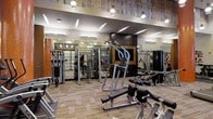 This single-floor fitness center is thoughtfully designed to accommodate a comprehensive workout experience. The space features a generous open floor plan filled with a diverse selection of strength training machines, including cable systems, lat pulldown stations, chest press units, and various selectorized equipment arranged to allow comfortable circulation throughout the facility.

The interior design blends industrial and modern aesthetics, highlighted by a textured wood-panel ceiling treatment that adds warmth and visual interest overhead. Recessed and track lighting fixtures provide bright, even illumination across the entire space, ensuring excellent visibility during workouts. Large floor-to-ceiling mirrors line the walls, enhancing the sense of depth and allowing users to monitor their form effectively.

The flooring consists of patterned, multi-toned carpet tiles that provide durability and comfort underfoot. The layout is well-organized, with equipment spaced generously to prevent congestion. The overall ambiance is professional and inviting, balancing functionality with a polished aesthetic that reflects a high-quality fitness facility suitable for users of varying fitness levels.