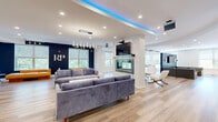 Located at 1 Richmond St in New Brunswick, NJ, this contemporary community space delivers a sophisticated and energetic atmosphere across a spacious single-floor layout. The interior is unified by light hardwood flooring, recessed lighting, and a crisp white-and-navy color palette that lends a clean, modern aesthetic throughout.

The main living area serves as the heart of the space, anchored by a stunning double-sided glass fireplace set within a textured stone column. Ambient blue LED cove lighting traces the curved ceiling above, while plush gray sofas, a live-edge coffee table, and a wall-mounted TV create an inviting lounge environment. An adjacent seating zone features tan leather daybeds positioned beneath large windows framing lush outdoor greenery, further brightening the space with natural light.

The second living area functions as a game and recreation room, highlighted by a pool table beneath a striking ceiling design, a shuffleboard table, and additional lounge seating — all bathed in natural light from multiple windows.

The kitchen is sleek and well-appointed, featuring white cabinetry, white quartz countertops, a textured glass peninsula with bar seating, pendant lighting, and stainless steel appliances including a built-in microwave. A row of glass-top high tables lines the window wall of the hallway, creating an ideal workspace or casual dining area illuminated by natural light and elegant chandeliers.
