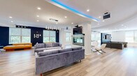 Located at 1 Richmond St in New Brunswick, NJ, this contemporary community space delivers a sophisticated and energetic atmosphere across a spacious single-floor layout. The interior is unified by light hardwood flooring, recessed lighting, and a crisp white-and-navy color palette that lends a clean, modern aesthetic throughout.

The main living area serves as the heart of the space, anchored by a stunning double-sided glass fireplace set within a textured stone column. Ambient blue LED cove lighting traces the curved ceiling above, while plush gray sofas, a live-edge coffee table, and a wall-mounted TV create an inviting lounge environment. An adjacent seating zone features tan leather daybeds positioned beneath large windows framing lush outdoor greenery, further brightening the space with natural light.

The second living area functions as a game and recreation room, highlighted by a pool table beneath a striking ceiling design, a shuffleboard table, and additional lounge seating — all bathed in natural light from multiple windows.

The kitchen is sleek and well-appointed, featuring white cabinetry, white quartz countertops, a textured glass peninsula with bar seating, pendant lighting, and stainless steel appliances including a built-in microwave. A row of glass-top high tables lines the window wall of the hallway, creating an ideal workspace or casual dining area illuminated by natural light and elegant chandeliers.