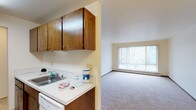 This single-floor unit is designed with a practical, open layout that connects the kitchen and living area seamlessly. The kitchen features warm wood-toned upper cabinetry with simple hardware, providing ample storage space. Light-colored countertops complement the cabinetry, and a stainless steel sink sits beneath a window that brings in natural light. White appliances complete the kitchen's functional setup.

Adjacent to the kitchen, the living area is finished with neutral wall-to-wall carpeting that adds a sense of warmth and comfort to the space. A large sliding glass door or picture window dominates the far wall, flooding the room with natural light and offering views of the surrounding greenery outdoors. Vertical blinds provide privacy and light control as needed. A baseboard heating unit runs along the base of the exterior wall, ensuring efficient climate control throughout the space. The overall color palette of warm neutrals and natural wood tones creates a cohesive, understated ambiance throughout the unit.