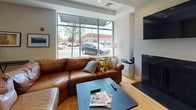 Located at 30 Haven St in Reading, MA, this single-floor community space offers a thoughtfully designed open-plan layout that seamlessly connects multiple functional areas. The flooring throughout is warm-toned hardwood, lending a cohesive and inviting atmosphere across the entire space.

The lounge area is anchored by a generous leather sectional sofa, a wall-mounted flat-screen television, and a fireplace, with large floor-to-ceiling windows providing abundant natural light and direct views of the surrounding streetscape. Framed artwork and accent tables add a curated, comfortable feel to this gathering zone.

Adjacent to the lounge is a dedicated dining area, furnished with a solid wood table and mission-style chairs that seat six. Glass partition walls and sliding doors visually connect this area to the entry corridor, maintaining an open and airy sense of flow. Built-in cabinetry with dark countertops along one wall offers additional storage and serving space.

Two distinct kitchen areas serve the space. One features a U-shaped layout with light maple cabinetry, dark granite countertops, a deep sink, and appliances including a microwave and range. The second is a more compact kitchen with an island, bar seating, stainless steel refrigerator, and additional appliances — ideal for communal use. Recessed lighting throughout ensures the space remains bright and functional at all times.