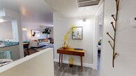This single-floor residence showcases a cohesive mid-century modern aesthetic carried throughout every room. The entryway sets an inviting tone with wood-look flooring, a warm accent console table, and curated wall art, offering a glimpse into both the living area and bedroom beyond.

The living room is a standout space, filled with natural light filtered through sheer curtains. A plush light-gray sofa anchored by a colorful striped area rug sits alongside a mustard accent chair, a tall fiddle-leaf fig, and an arching brass floor lamp — creating a lively yet comfortable atmosphere.

The dining area is defined by a round wood table surrounded by teal velvet chairs beneath a sleek chandelier, with a pass-through connecting it to the kitchen. The kitchen features white cabinetry with brushed hardware, black appliances including an electric range and refrigerator, and granite-style countertops, offering a practical and clean workspace.

The bedroom provides a calm retreat with wood-look flooring, a natural light-filled window with vertical blinds, a built-in closet with shelving, and soft wall sconces. The full bathroom includes a tub-shower combination, a vanity with ample counter space, and white cabinetry, finished with cheerful accent décor throughout.