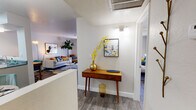 This single-floor residence showcases a cohesive mid-century modern aesthetic carried throughout every room. The entryway sets an inviting tone with wood-look flooring, a warm accent console table, and curated wall art, offering a glimpse into both the living area and bedroom beyond.

The living room is a standout space, filled with natural light filtered through sheer curtains. A plush light-gray sofa anchored by a colorful striped area rug sits alongside a mustard accent chair, a tall fiddle-leaf fig, and an arching brass floor lamp — creating a lively yet comfortable atmosphere.

The dining area is defined by a round wood table surrounded by teal velvet chairs beneath a sleek chandelier, with a pass-through connecting it to the kitchen. The kitchen features white cabinetry with brushed hardware, black appliances including an electric range and refrigerator, and granite-style countertops, offering a practical and clean workspace.

The bedroom provides a calm retreat with wood-look flooring, a natural light-filled window with vertical blinds, a built-in closet with shelving, and soft wall sconces. The full bathroom includes a tub-shower combination, a vanity with ample counter space, and white cabinetry, finished with cheerful accent décor throughout.