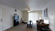 This single-floor residence offers a warm and functional layout designed for comfortable everyday living. The living room serves as the heart of the home, featuring soft wall-to-wall carpeting that adds a sense of coziness underfoot. A ceiling fan with an integrated light fixture is centrally mounted, providing both air circulation and ambient lighting throughout the space. A large sliding glass door dressed with vertical blinds fills the room with natural light while offering the option for privacy, creating a bright and airy atmosphere. The living area is well-proportioned, providing ample room for a full seating arrangement, including a plush sofa and a rocking chair with an ottoman — ideal for relaxing. A dark-finished TV console anchors one side of the room, complementing the dark wood coffee table at the center. Neutral-toned walls and a clean white ceiling contribute to a calm, understated aesthetic that allows for flexible interior styling. A floor lamp in the corner adds warm accent lighting, enhancing the overall inviting ambiance. The space is thoughtfully arranged to balance function and comfort, making it a welcoming environment for both everyday use and casual entertaining.