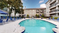 This outdoor amenity space centers around a generously sized freeform swimming pool with clear, blue-tinted water and a smooth concrete surround. The pool deck is well-appointed with numerous blue sling-style lounge chairs and side tables, offering ample space for sunbathing and relaxation. Blue market umbrellas are positioned throughout the deck, providing shaded retreats on sunny days, while festoon string lights are strung between mature trees to create an inviting ambiance during evening hours. Large cobalt-blue planters filled with lush greenery serve as decorative accents, adding vibrant color throughout the space. A dedicated outdoor dining area features umbrella-shaded tables with chairs, ideal for casual gatherings. The courtyard is enclosed by a black iron fence and framed by mature magnolia and shade trees that contribute to a lush, park-like atmosphere. The surrounding multi-story building, finished in a warm gray tone with arched window detailing, classical moldings, and private balconies, creates a cohesive architectural backdrop. The overall design of the courtyard balances aesthetic appeal with functional amenities, making it a central social and recreational hub for the community.