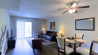 This two-floor residence offers a thoughtfully designed layout that combines comfort and functionality. The main living and dining area showcases rich hardwood flooring that extends throughout the open-concept space, creating a warm and cohesive aesthetic. A ceiling fan with integrated lighting fixture is centrally positioned overhead, providing both air circulation and ambient illumination.

The living area is spacious enough to accommodate a full seating arrangement and features large sliding glass doors dressed with vertical blinds, allowing abundant natural light to filter in while offering direct access to a private outdoor balcony. The balcony extends the living space outdoors and provides an additional area for relaxation.

Adjacent to the living room, the dining area comfortably fits a full dining table and chair set, making it well-suited for everyday meals and entertaining. The open flow between these two zones enhances the sense of space and encourages easy movement throughout.

The walls are finished in a neutral tone, complemented by a textured ceiling that adds subtle visual interest. Warm accent lighting from table lamps contributes to an inviting atmosphere throughout the shared living spaces. The overall design prioritizes an open, airy feel across both floors of the home.