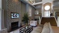 This impressive home showcases a dramatic, open-concept living area defined by soaring two-story ceilings with exposed white wood beam detailing, creating an immediate sense of grandeur upon entry. The focal point of the main living space is a floor-to-ceiling stacked stone fireplace, flanked by a wall-mounted flat-screen television, adding both warmth and visual impact to the room. Neutral, earthy tones across the walls complement the rich hardwood flooring and plush, earth-toned upholstered furnishings, including a tufted accent chair and a glass-top coffee table that anchor the seating area. A tall indoor palm plant introduces a natural, organic element to the space. Natural light filters in through multiple windows, including a distinctive round porthole-style window near the upper level, enhancing the airy atmosphere. A handsome staircase with white-painted newel posts and dark wood railings leads to an upper landing, visible from the main living area. The architectural design blends traditional and transitional styles, with coffered ceiling elements and clean white trim providing a refined finish throughout. The overall ambiance is welcoming, sophisticated, and well-suited for both everyday living and entertaining.