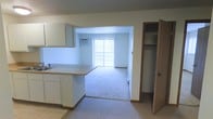 This single-floor space offers a practical and straightforward layout designed for comfortable everyday living. The kitchen is outfitted with white cabinetry and a light-toned countertop that extends as a peninsula, creating a natural divide between the kitchen and the adjacent living area. The flooring transitions from hard surface in the kitchen to plush carpet in the living space, adding warmth and comfort underfoot. A large sliding glass door anchors the living area, flooding the space with natural light through vertical blinds and providing access to the outdoors. A wall-mounted air conditioning unit ensures year-round climate control. Just off the main living area, a built-in closet with a hanging rod offers organized storage space. The overall color palette is neutral and clean, with white walls and warm wood-toned trim accents throughout. The open flow between the kitchen and living area creates a cohesive, functional environment well-suited for day-to-day use. The single-floor layout ensures easy accessibility and a straightforward living experience.