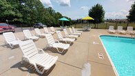 This outdoor swimming pool area centers around a generously sized freeform pool with clearly marked depth indicators ranging from shallow to deep ends, complemented by built-in metal entry rails for safe access. The pool features clean, blue water and a smooth tiled border, set within a wide concrete sundeck that provides ample space for relaxation and circulation.

The sundeck is furnished with numerous white lounge chairs arranged throughout the pool surround, along with shaded seating areas equipped with colorful market umbrellas and outdoor table-and-chair sets, ideal for socializing or dining poolside. The entire pool area is enclosed by a black metal fence, ensuring safety and security.

Adjacent to the pool deck stands a brick and Tudor-style building that houses the pool's support facilities. Inside, dedicated utility rooms contain the pool's mechanical infrastructure, including a filtration system with visible PVC piping and pump equipment. Additional storage rooms feature floor-to-ceiling shelving stocked with pool maintenance chemicals, cleaning supplies, and general maintenance materials.

The surrounding landscape includes mature trees that provide a natural backdrop and partial shade, enhancing the outdoor ambiance. The overall setting offers a well-maintained, functional, and inviting outdoor recreational environment.