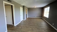 This single-floor space offers a clean, modern aesthetic characterized by a cohesive neutral gray color palette on the walls, crisp white baseboards, and warm wood-look laminate flooring throughout. The main room is generously proportioned, providing an open and versatile layout that can accommodate a range of furniture configurations and uses. The textured popcorn ceiling adds a classic touch, while flush-mount ceiling lighting ensures consistent illumination throughout the space. Natural light filters in through a window along one wall, brightening the interior and enhancing the overall sense of openness. Multiple doorways lead to adjoining areas, including what appears to be a closet or storage space and a secondary room with warm-toned walls visible through the doorway, suggesting additional functional square footage. The overall design is straightforward and practical, with a move-in-ready feel. The combination of durable flooring, neutral tones, and well-defined spaces makes this property highly adaptable for residential living. The clean lines and consistent finishes throughout create a unified, inviting atmosphere that serves as an excellent blank canvas for personalization.