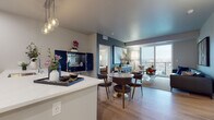 This well-appointed single-floor residence showcases a thoughtfully designed open-concept layout that seamlessly connects the kitchen, dining, and living areas. The kitchen is outfitted with white shaker cabinetry, stainless steel appliances including a refrigerator, range, and microwave, a herringbone tile backsplash, and a generously sized island with quartz countertops and pendant lighting overhead. Light wood-style flooring runs throughout the main living areas, contributing to a bright and cohesive aesthetic.

The dining area accommodates a round table with modern seating, positioned adjacent to a statement navy cabinet that doubles as a bar. The living room features large floor-to-ceiling sliding glass doors that open to a private balcony, offering expansive city views and abundant natural light. Bold accent walls and curated artwork add visual depth to the space.

Both bedrooms are carpeted and finished with neutral tones, providing a calm retreat. Each room includes windows with city views and ample space for furnishings. The bathroom features light wood-style flooring, white cabinetry with a quartz vanity top, and a clean, minimalist design. A dedicated entryway with a navy front door completes the unit.