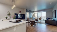 This well-appointed single-floor residence showcases a thoughtfully designed open-concept layout that seamlessly connects the kitchen, dining, and living areas. The kitchen is outfitted with white shaker cabinetry, stainless steel appliances including a refrigerator, range, and microwave, a herringbone tile backsplash, and a generously sized island with quartz countertops and pendant lighting overhead. Light wood-style flooring runs throughout the main living areas, contributing to a bright and cohesive aesthetic.

The dining area accommodates a round table with modern seating, positioned adjacent to a statement navy cabinet that doubles as a bar. The living room features large floor-to-ceiling sliding glass doors that open to a private balcony, offering expansive city views and abundant natural light. Bold accent walls and curated artwork add visual depth to the space.

Both bedrooms are carpeted and finished with neutral tones, providing a calm retreat. Each room includes windows with city views and ample space for furnishings. The bathroom features light wood-style flooring, white cabinetry with a quartz vanity top, and a clean, minimalist design. A dedicated entryway with a navy front door completes the unit.