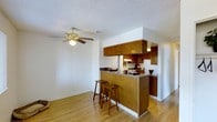 This single-floor space showcases an open-concept layout that seamlessly connects the kitchen and living area, creating a functional and cohesive environment. The kitchen is outfitted with warm medium-toned wood cabinetry extending from the lower counters to the upper wall-mounted cabinets, providing generous storage. A peninsula-style breakfast bar with a dark countertop serves as both a prep space and a casual dining area, accompanied by two wooden barstools. Standard appliances, including a refrigerator and stovetop range with an overhead vent hood, are neatly integrated into the kitchen layout. Light hardwood flooring runs throughout the space, adding warmth and continuity between the kitchen and the adjoining living area. The living area is open and uncluttered, benefiting from a ceiling fan with a built-in light fixture that promotes air circulation and ambient lighting. The textured popcorn ceiling and white walls provide a neutral backdrop, while the open sightlines between rooms enhance the sense of spaciousness. Overall, the space offers a practical and comfortable layout suited for everyday living.