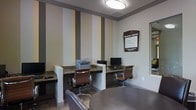 This Business Center is a single-floor workspace designed to meet the professional needs of its users. The room features multiple built-in workstations with granite-style countertops, each equipped with a desktop computer monitor, providing dedicated spaces for individual work. A multifunction printer sits conveniently along the counter, supporting printing and copying needs. The seating consists of ergonomic brown leather office chairs, ensuring comfort during extended work sessions. A round table with additional seating is positioned in the foreground, offering a collaborative or meeting area. The walls are decorated with bold vertical stripes in neutral tones of gray, tan, and cream, adding visual interest and a modern aesthetic. Flush-mounted ceiling lighting provides consistent, even illumination throughout the space. A clearly posted Business Center sign outlines usage guidelines for occupants. A large mirror or window on the right wall enhances the sense of openness. Overall, the space maintains a clean, professional ambiance well-suited for business tasks.