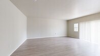 This studio presents a clean, functional layout making efficient use of its single-floor design. The open living area is bright and airy, featuring light gray wood-style flooring and neutral white walls that create a fresh, minimalist atmosphere. Natural light filters in through multiple windows fitted with blinds, including a large sliding door covered by vertical blinds that further enhances the sense of openness. The kitchen is thoughtfully appointed with light maple cabinetry, gray laminate countertops, a double-basin stainless steel sink, and black appliances including an electric range, built-in microwave, refrigerator, and dishwasher. The L-shaped counter layout maximizes workspace and storage efficiency. The bathroom features light wood-toned cabinetry with a dark countertop vanity, a mirror, built-in open shelving, a full-size soaking tub with a shower and curtain rod, and the same wood-style flooring carried throughout the unit. The space also includes a dedicated closet for additional storage. The consistent use of neutral tones and light flooring throughout ties the entire studio together, creating a cohesive and inviting environment.