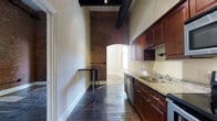 This single-floor residence blends historic architectural charm with modern functionality across a thoughtfully designed layout featuring one bedroom, one bathroom, and a versatile den. The space immediately impresses with its soaring ceilings, which lend an open, airy quality throughout. Exposed brick columns and archways serve as striking focal points, adding raw, industrial character that contrasts beautifully with the clean white walls and dark, rich hardwood floors underfoot. Visible ductwork along the ceiling further reinforces the loft-inspired aesthetic.

The galley-style kitchen is well-equipped and efficiently arranged, featuring rich dark wood cabinetry, granite countertops, and stainless steel appliances, including a dishwasher. A double sink with modern fixtures completes the space. Natural light filters in through a window at the end of the kitchen corridor, brightening the area. A narrow wall-mounted table provides a convenient spot for casual dining or workspace just off the kitchen. The arched brick passageway leads seamlessly between areas, enhancing the sense of historic character that defines this distinctive residence.
