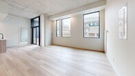 This contemporary 2-bedroom, 2-bathroom unit is designed with a clean, modern aesthetic throughout. The open-concept living and dining area is a standout space, featuring striking exposed concrete ceilings, light-toned hardwood flooring, and oversized black-framed windows that flood the interior with natural light and provide urban views of the surrounding cityscape. A sliding glass door extends the living area toward an outdoor access point, enhancing the sense of openness.

The kitchen is seamlessly integrated into the main living space and features a central island with a light wood finish, white gloss upper cabinetry, warm wood-toned lower cabinets, and stainless steel appliances, creating a functional yet stylish workspace.

Both bedrooms carry the same light hardwood flooring and crisp white walls, with exposed concrete accents adding an industrial touch. Each room benefits from natural light via well-placed windows with urban outlooks. The bedrooms are accessed via a central hallway with clean white paneled doors featuring matte black hardware.

The two bathrooms are finished in a minimalist style, with white cabinetry, white quartz countertops, undermount sinks, and terrazzo-style tile flooring. One bathroom includes a full soaking tub with subway tile surround, while both feature sleek rectangular mirrors above the vanities.