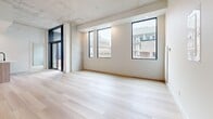 This contemporary 2-bedroom, 2-bathroom unit is designed with a clean, modern aesthetic throughout. The open-concept living and dining area is a standout space, featuring striking exposed concrete ceilings, light-toned hardwood flooring, and oversized black-framed windows that flood the interior with natural light and provide urban views of the surrounding cityscape. A sliding glass door extends the living area toward an outdoor access point, enhancing the sense of openness.

The kitchen is seamlessly integrated into the main living space and features a central island with a light wood finish, white gloss upper cabinetry, warm wood-toned lower cabinets, and stainless steel appliances, creating a functional yet stylish workspace.

Both bedrooms carry the same light hardwood flooring and crisp white walls, with exposed concrete accents adding an industrial touch. Each room benefits from natural light via well-placed windows with urban outlooks. The bedrooms are accessed via a central hallway with clean white paneled doors featuring matte black hardware.

The two bathrooms are finished in a minimalist style, with white cabinetry, white quartz countertops, undermount sinks, and terrazzo-style tile flooring. One bathroom includes a full soaking tub with subway tile surround, while both feature sleek rectangular mirrors above the vanities.