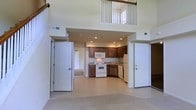 This two-story residence is defined by its dramatic double-height ceilings in the main living area, which create an expansive, open atmosphere and allow natural light to flow throughout the space. An upper-level balcony with a white railing overlooks the main floor, adding architectural interest and reinforcing the sense of vertical space.

The open-concept layout seamlessly connects the living area to the kitchen, which features warm medium-toned wood cabinetry arranged in an efficient L-shaped configuration. The kitchen is equipped with white appliances, including a range, built-in microwave, dishwasher, and refrigerator, as well as a sink positioned along the back counter. Recessed lighting illuminates the kitchen area, complementing the natural light from above.

The main living space is finished with light-toned carpet, while the kitchen transitions to tile flooring for a practical and clean distinction between areas. Two interior doors are visible on either side of the main space, providing access to additional rooms. The overall palette is neutral and bright, with white walls and trim contributing to the home's clean, airy character.